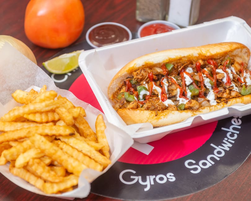 A chicken cheesesteak sandwich with fries, ketchup, BBQ sauce, a tomato, onion, and lime.